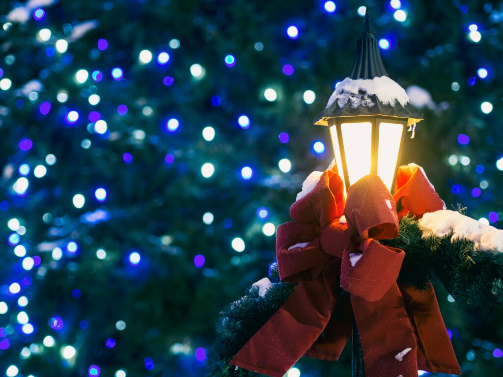 A streetlamp has a red bow and some snow on it, while the background looks like a tree with white and blue lights in it. Enjoy the holiday season and get our cleaning services Sugar Land.