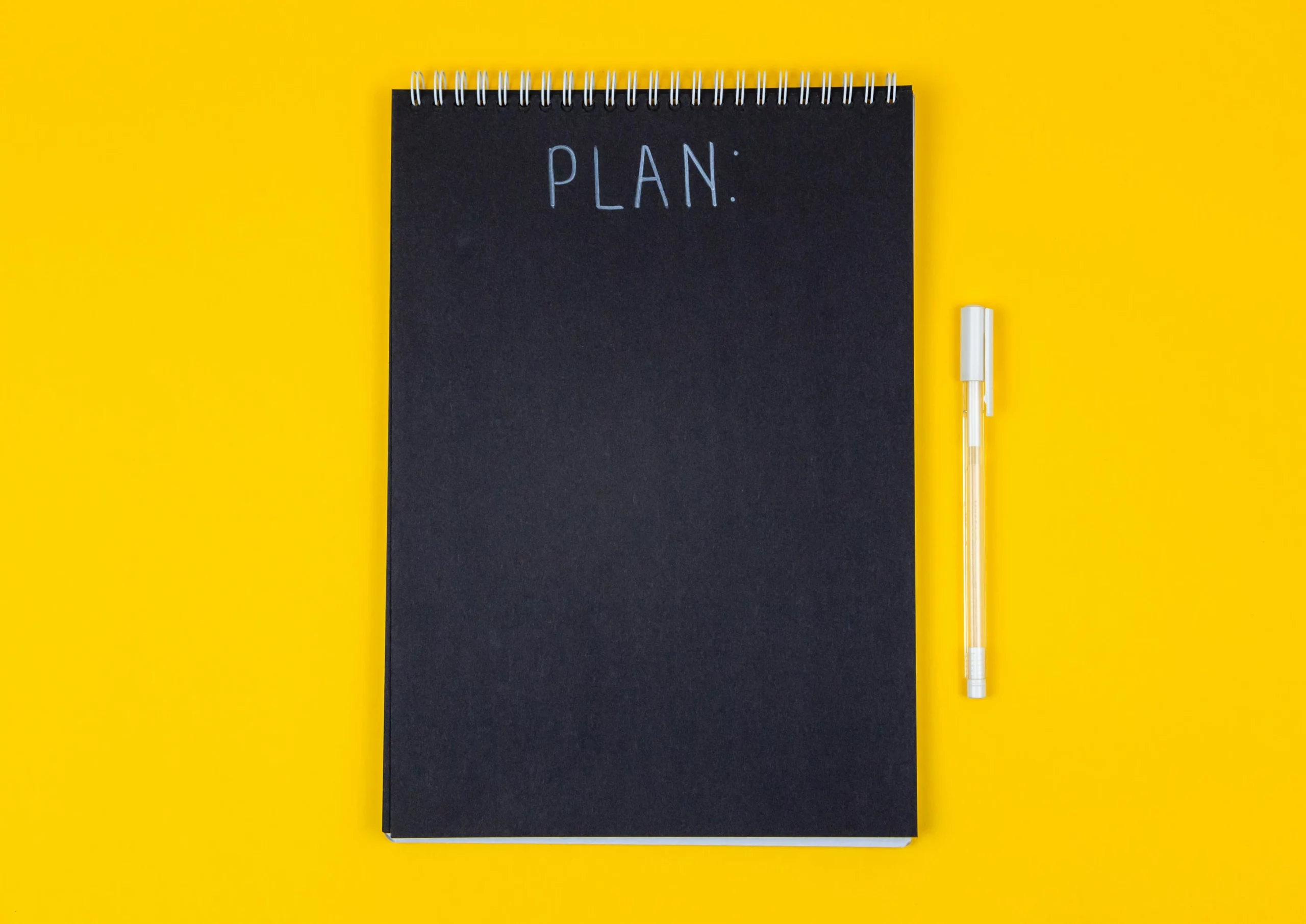 A black notebook that says plan at the top, with a white pen beside it, above a yellow background. Somone is going to write a plan for ADHD cleaning tips.