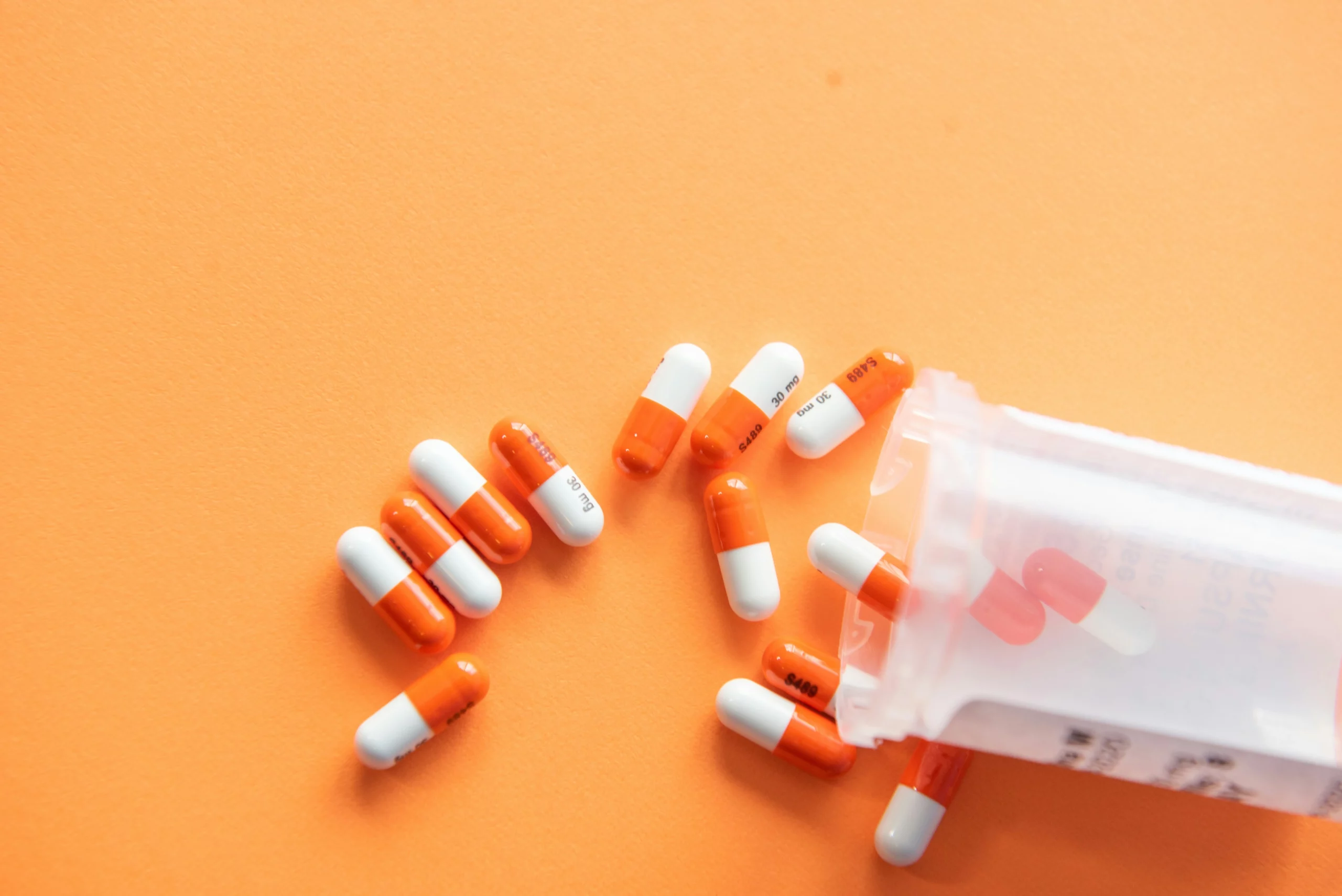 Medicine capsules pour out of a medicine bottle over an orange background. People who take ADHD meds may need to pair that treatment with ADHD cleaning tips.