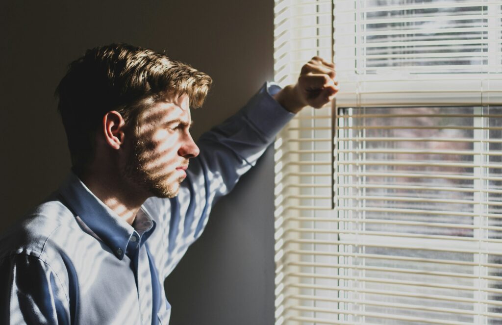 A man is in a dark room looking out a window during the day. He is overwhelmed and depressed, but he has to figure out how to clean his house.