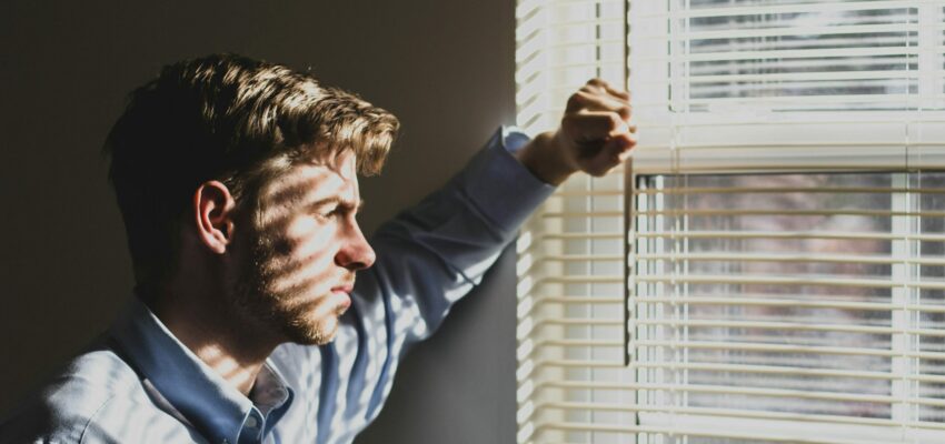 A man is in a dark room looking out a window during the day. He is overwhelmed and depressed, but he has to figure out how to clean his house.