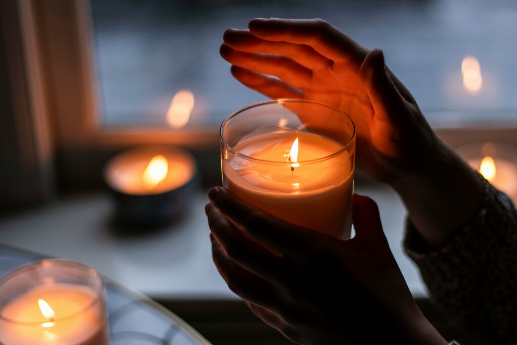 Someone is holding a lit candle in their hands, in a glass jar. There are other lit candles in the background.