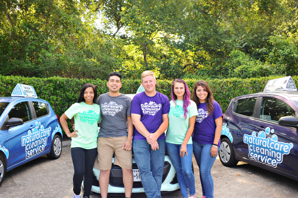 A group of maids from Naturalcare Cleaning Service are posing for a photo in front of their branded vehicles, in a parking lot on a summer day. They are cleaners Rosenberg TX sees often.