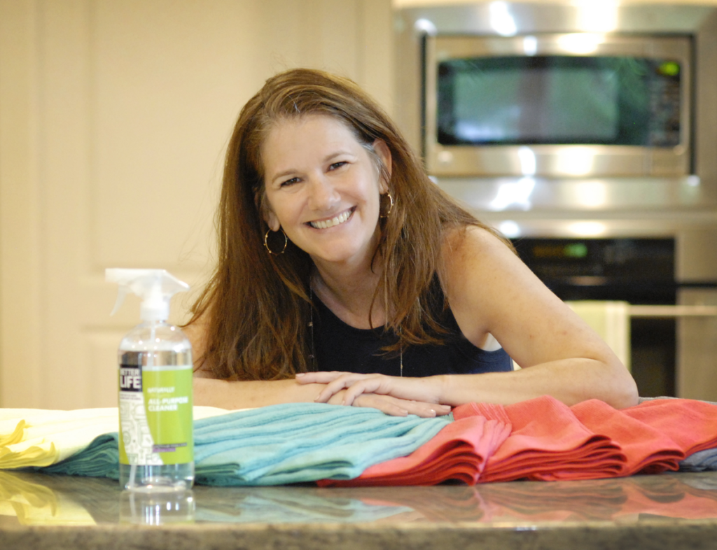 Eco friendly supplies are on a counter. A woman from Naturalcare Cleaning Services is smiling at the camera, over a kitchen counter.